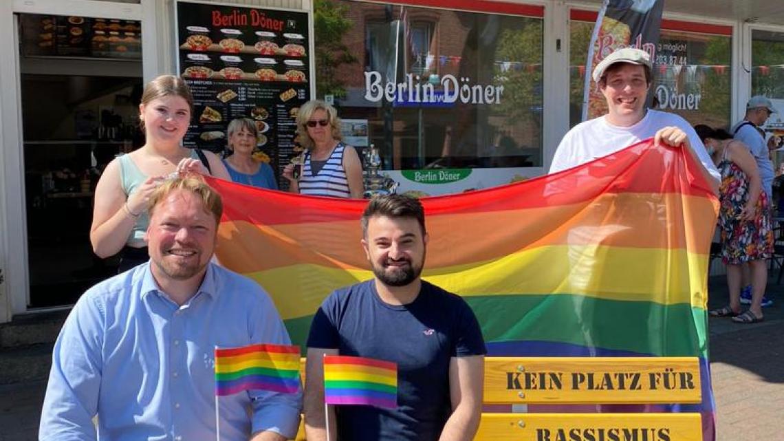 Eine Schülerin und ein Schüler der Gemeinschaftsschule Brunsbüttel (=Schule ohne Rassismus) halten die Regenbogenflagge. Auf der Bank links: Oliver Kumbartzky. Rechts daneben Delil Uca.